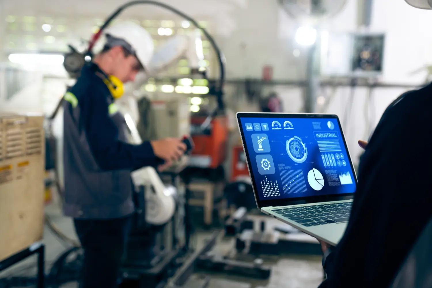 Techniker überwacht industriellen Roboterarm per Laptop mit digitalem Steuerungs-Dashboard in einer Produktionshalle