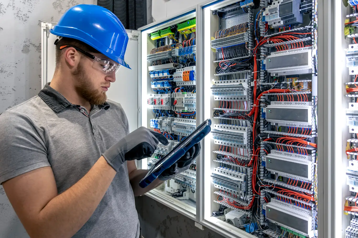 Elektrotechniker mit Schutzhelm und Tablet prüft verdrahteten Schaltschrank bei der KNX-Programmierung