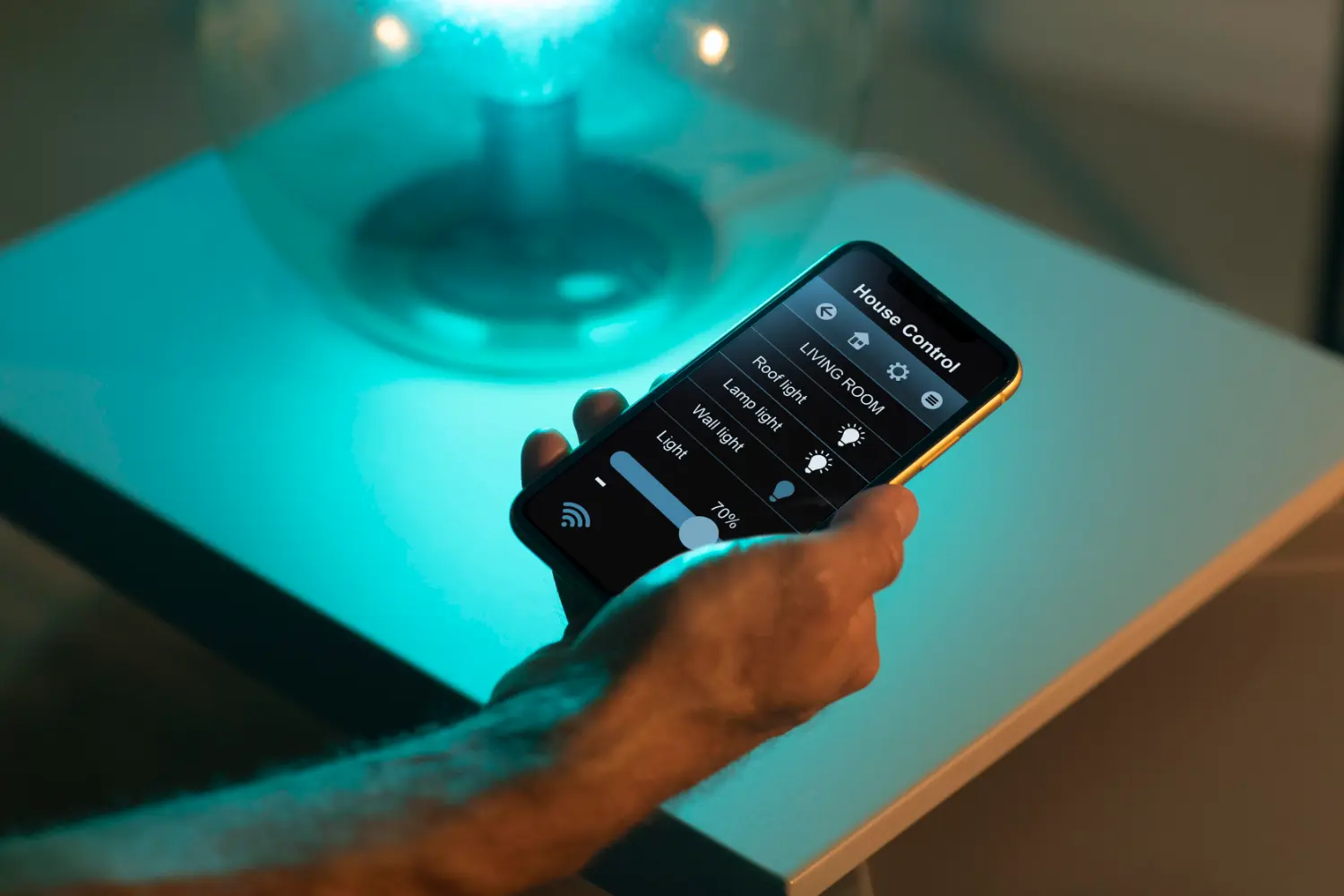 Hand hält Smartphone mit Smart-Home-App zur Lichtsteuerung im Wohnzimmer bei gedimmtem blauen Licht